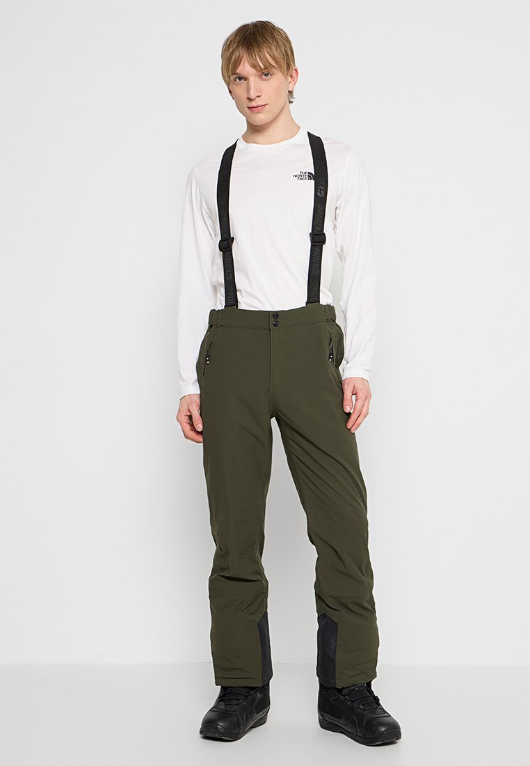 Junger Mann in einem weißen Langarmhemd, olivgrünen Schneehosen mit schwarzen Trägern und schwarzen Stiefeln, vor einem schlichten Hintergrund stehend.