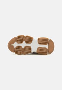 Semelle de chaussure avec un motif texturé en caoutchouc brun clair, présentant des rainures en zigzag. La semelle intermédiaire blanche se connecte à la partie supérieure.