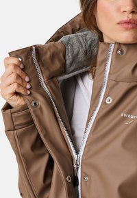 Brun vattentålig jacka med en mjuk grå fleecefoder, zippered fram, och tryckknappar. Har logotyp och kontrasterande sömmar.