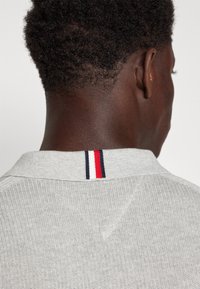 Grå stickad pikétröja med en minimal krage som har röda, vita och marinblå ränder. Närbild av baksidan av halsringningen och texturen.