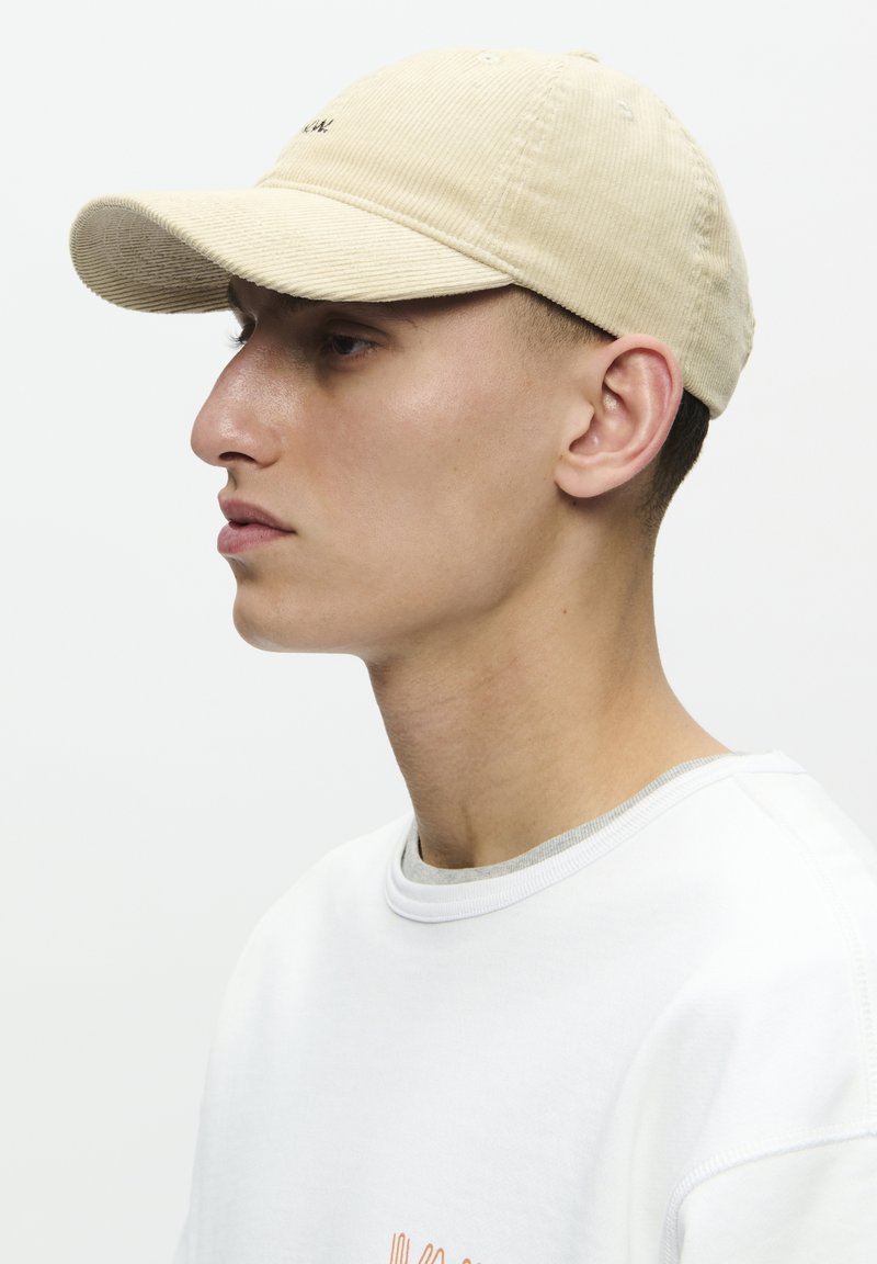 Beige corduroy baseball cap with a curved brim, featuring a small embroidered logo on the front. Styled with a white shirt.