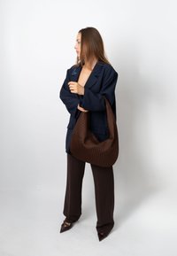 Pruun punutud boho-kott tekstuurse pinnaga, kombineeritud tumelilla oversize bleiseri ja laiade pruunide pükstega, valge taustaga.