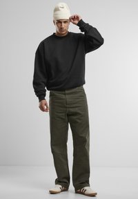 Felpa nera, pantaloni larghi verde oliva e berretto di lana bianco. Le calzature presentano una base crema con strisce marroni. Elementi di design minimalisti.
