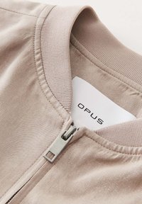 Lichtbruine ritsjack met een gladde textuur en een geribbelde kraag. Beschikt over een zilveren rits en een label met "OPUS."