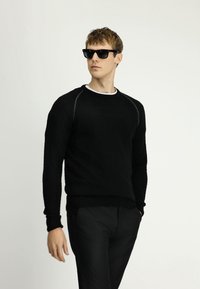 Czarny sweter o teksturowanym wzorze z okrągłym dekoltem, ściągaczami przy mankietach i u dołu. Noszony z czarnymi okularami przeciwsłonecznymi i czarnymi spodniami, tworząc monochromatyczny look.