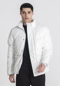 Hvid pufferjakke med quiltet design, høj krave, frontlynlås og sorte trykknapper. Elegant glansfuld finish og afslappet pasform.