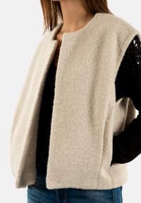 Gilet di lana beige con finitura testurizzata, davanti aperto e tasche laterali senza cuciture, indossato sopra una maglia nera. Senza chiusure.