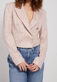 Blazer doppiopetto in tweed rosa chiaro con ampi revers, tessuto strutturato e bottoni metallici, abbinato a jeans in denim blu a vita alta.