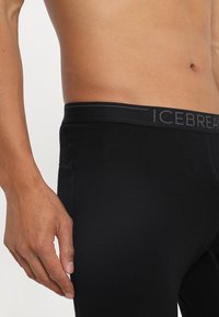 Leggings thermiques noirs avec une texture lisse, dotés d'une large taille étiquetée "ICEBREAKER" en gris. Gros plan montrant la main droite sur la hanche.