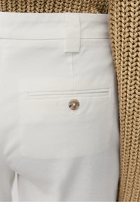 Un pantalon en coton blanc présente une poche arrière avec un bouton rond beige. Le tissu a une texture lisse, tandis que la taille est plate.