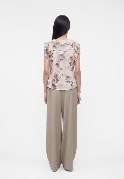 Γυναίκα με μακριά μαύρα μαλλιά που φοράει αμάνικη φλοράλ μπλούζα με βολάν στα μανίκια και μπεζ παντελόνι με φαρδιά πόδια, με την πλάτη σε λευκό φόντο.