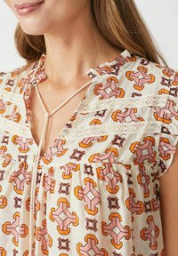 Top a maniche corte in tessuto leggero con motivo floreale nei colori rosa, arancione e marrone. Presenta dettagli in pizzo e un nodo al collo.