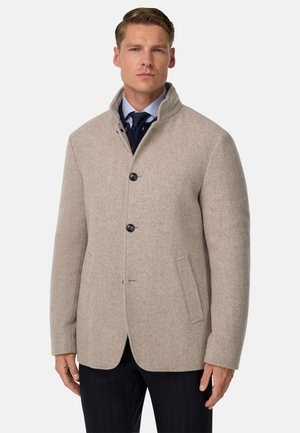 Beige Wollmischungsjacke mit hohem Kragen, zwei Vorderaschen, drei Knöpfen und glatter Textur, kombiniert mit einem marineblauen gemusterten Hemd.