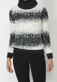 Eine Person trägt einen samtigen schwarz-weiß gestreiften Rollkragenpullover mit langen Ärmeln und schwarze Hosen und steht vor einem schlichten Hintergrund.