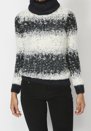 Persoon in een fuzzy zwart-wit gestreepte coltrui met lange mouwen en zwarte broek, staand tegen een effen achtergrond.