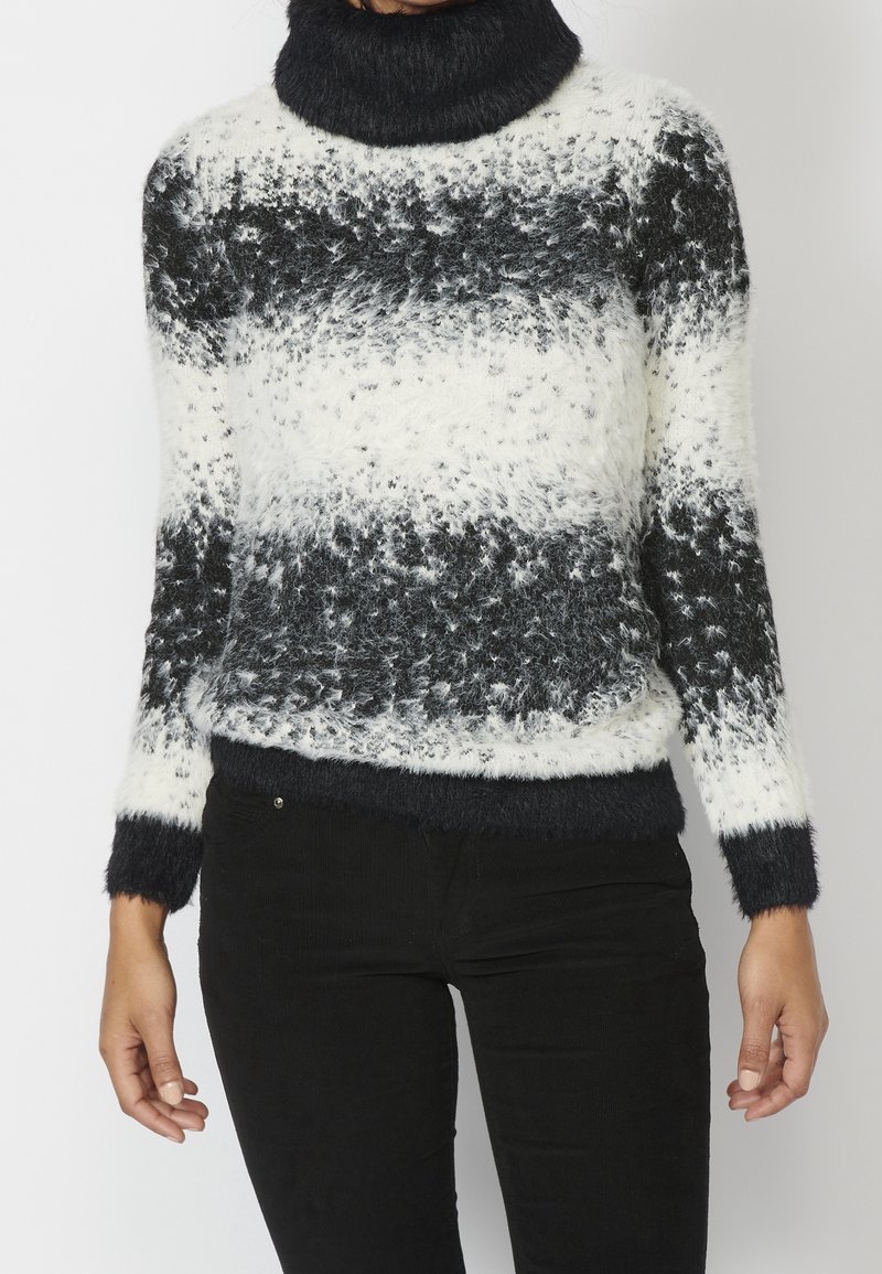 Eine Person trägt einen samtigen schwarz-weiß gestreiften Rollkragenpullover mit langen Ärmeln und schwarze Hosen und steht vor einem schlichten Hintergrund.