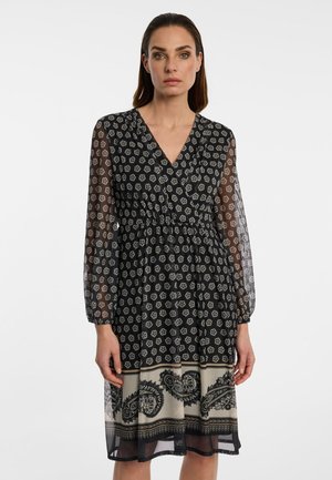 Woman wearing a black dress with white floral pattern, sheer long sleeves, V-neck, and a beige paisley print near the hem, standing against a white background.