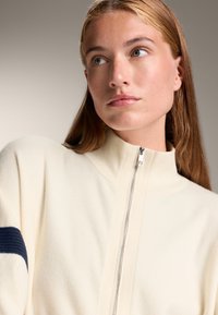 Cremfarbener Zip-Up-Pullover mit hohem Kragen, der einen dunkelblauen Streifen am linken Arm aufweist. Weicher Stoff und lockere Passform. Der Metallreißverschluss ist sichtbar.