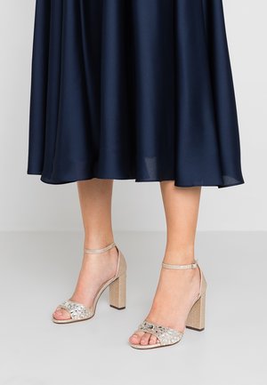 Sandalias de tacón bloque beige con un acabado texturizado, que cuentan con una correa adornada con strass. La correa suave para el tobillo añade estabilidad.