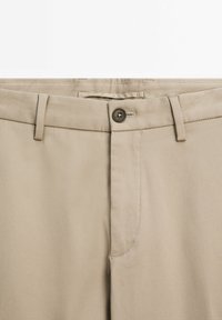 Beige trousers featuring a flat front, angled side pockets, a button closure, and subtle diagonal texture.