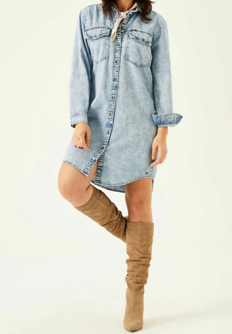 Denimklänning i ljusblått, med långa ärmar, två bröstfickor och knappstängning fram. Bärs med tanfärgade stövlar i mocka som går upp till knäna.