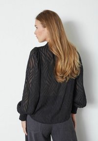 Blusa nera trasparente con motivo a onda, maniche lunghe, spalle arricciate e polsini texturizzati. Abbinata a pantaloni grigi, i capelli scendono oltre le spalle.