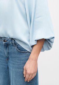 Άτομο που φοράει ανοιχτό μπλε μπλουζάκι με φαρδιά μανίκια και μπλε τζιν τζιν, το χέρι χαλαρό στο πλάι, ουδέτερο φόντο.