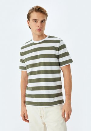 Camiseta de manga corta a rayas en verde oliva y blanco con cuello redondo, hecha de suave tejido de algodón. Modelo lleva puesto un pantalón de color claro.