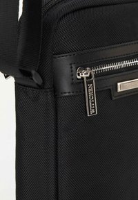 Borsa in tessuto nera con superficie testurizzata, dettagli in pelle, zip argentata e un'etichetta metallica con logo. Include una tracolla regolabile.