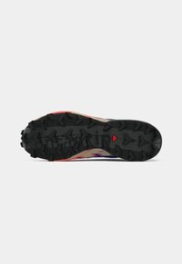Sola de sapatilha de trail running em borracha preta, com padrões profundos para aderência e uma mistura de detalhes em vermelho e roxo na parte superior.