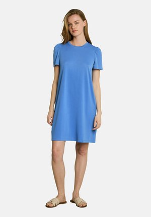 Woman standing wearing a knee-length, short-sleeve blue dress with puffed shoulders and beige flat sandals, against a plain light background.
