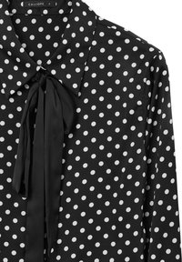 Schwarze Bluse mit weißen Pünktchen, Kragen mit Schleifen-Detail, lange Ärmel und glatte Stoffoberfläche. Markenlabel sichtbar.