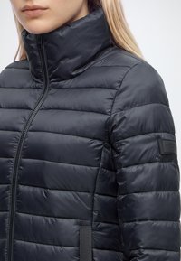 Femme portant une doudoune matelassée noire avec un col montant et une fermeture éclair visible, cadrée pour montrer la poitrine et les épaules.