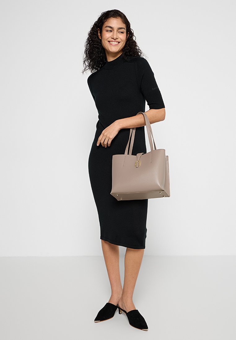 Braune Leder-Tote mit zwei Griffen, strukturierter Form und goldenen Hardware-Akzenten. Das Modell trägt ein schwarzes, tailliertes Kleid und schwarze Mules.