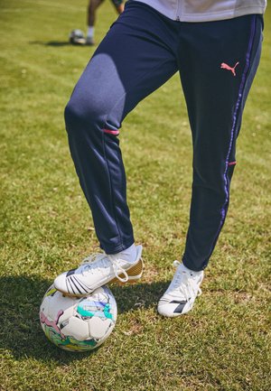 Tumedad spordipüksid roosa ja lilla aktsentidega, valgete jalgpallikingadega, puhkamas värvilise mustriga jalgpallil rohul.