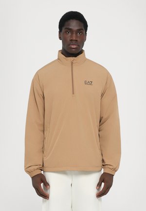 Tan half-zip jacket with a high collar, elastic cuffs, and branded logo. Made from lightweight fabric, featuring a relaxed fit.