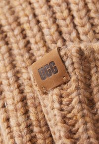 Tessuto a maglia marrone con un pattern intrecciato, dotato di un'etichetta in pelle tan rettangolare con la scritta "UGG" in rilievo. Superficie morbida e testurizzata.