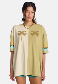 T-shirt oversize en coton avec sections divisées en beige et vert olive. Comprend une broderie de palmier et des accents décoratifs bleus et orange.