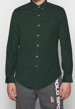 Man draagt een donkergroen overhemd met knopen en grijze Polo Sport joggingbroek met Amerikaanse vlag en merknaam op het been.