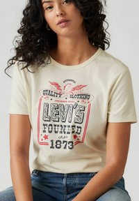 Beige katoenen T-shirt met een grafische print met rode en blauwe tekst, een adelaarontwerp en de tekst "Levi's Founded 1873" op de voorkant.