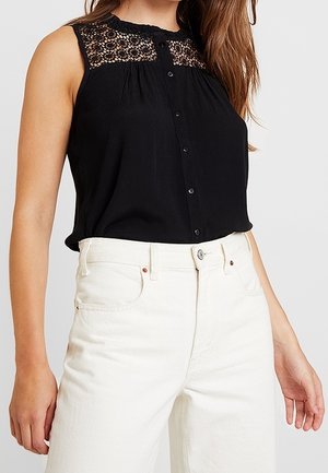 Femme portant un chemisier noir sans manches avec un empiècement en dentelle et un jean blanc taille haute, debout les bras relâchés le long du corps.