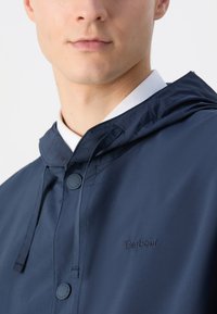 Navy blue hooded jacket with a smooth texture, featuring an embroidered logo, button closures, and drawstring detail at the neck.