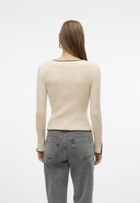 Top a maniche lunghe beige a coste con scollatura rotonda e rifiniture nere, abbinato a jeans grigi a vita alta con tasche posteriori.