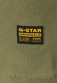 Svart och gul G-Star Originals Raw Denim tygmärke sytt på ribbad olivgrön textil.