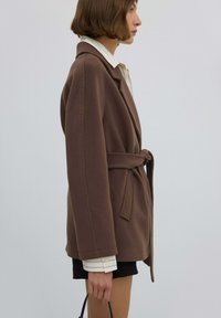 Brązowa kurtka z paskiem, kołnierzem z wycięciem, zapięciem na guziki, bocznymi kieszeniami i gładką fakturą. Ubrana na jasną koszulę i szorty.