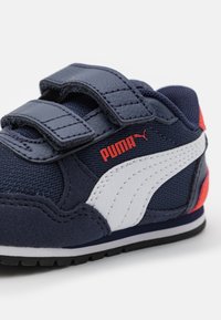 Námořnické modré a bílé tenisky Puma se dvěma suchými zipy, červeným logem Puma a červeným detailem na patě, zobrazené zblízka na bílém pozadí.