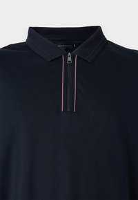 Marineblå pologenser med glidelås i kragen og to røde aksentstriper. Laget av et glatt stoff, med klassisk design og korte ermer.