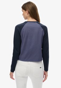 Kobieta z długimi brązowymi włosami, ubrana w niebieską bluzkę z długim rękawem typu raglan oraz białe spodnie, stojąca tyłem na tle jednolitego białego tła.