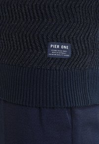 Pull en maille bleu marine foncé avec un motif en zigzag et une étiquette "Pier One" au-dessus d'un ourlet côtelé, associé à un pantalon texturé foncé.