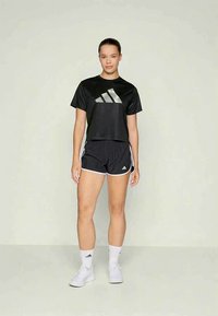 Noor noor seisev naine mustades Adidas spordirõivastes, valgetes sokkides ja valgetes tennisetossudes, lihtsa heleda tausta ees.